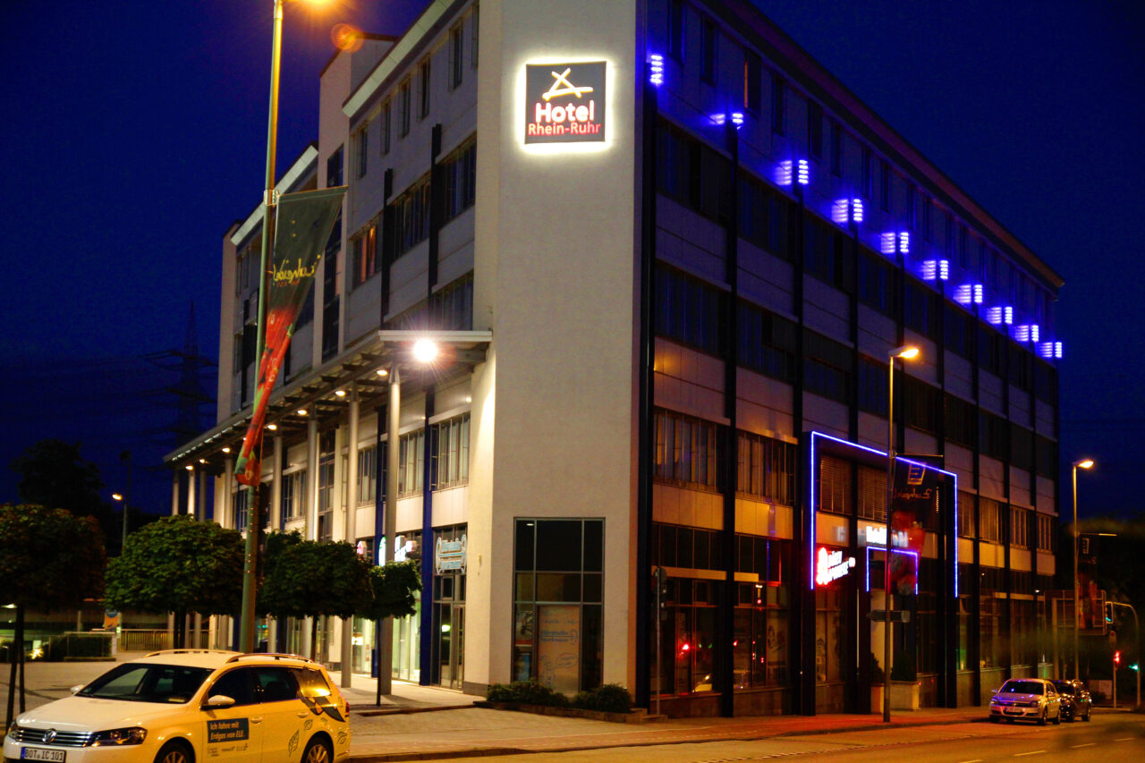 Rhein-Ruhr-Hotel, Bottrop: Beleuchtetes Hotelgebäude an einer Straße in Bottrop bei Nacht, mit Autos und blauem Neonlicht.