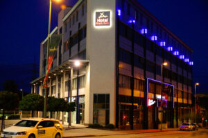 Rhein-Ruhr-Hotel, Bottrop: Beleuchtetes Hotelgebäude an einer Straße in Bottrop bei Nacht, mit Autos und blauem Neonlicht.