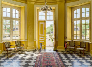 Schloß Beck, Bottrop: Heller Eingangsraum von Schloss Beck mit gelben Wänden, Kronleuchter, Marmorfliesen und Sitzmöbeln.