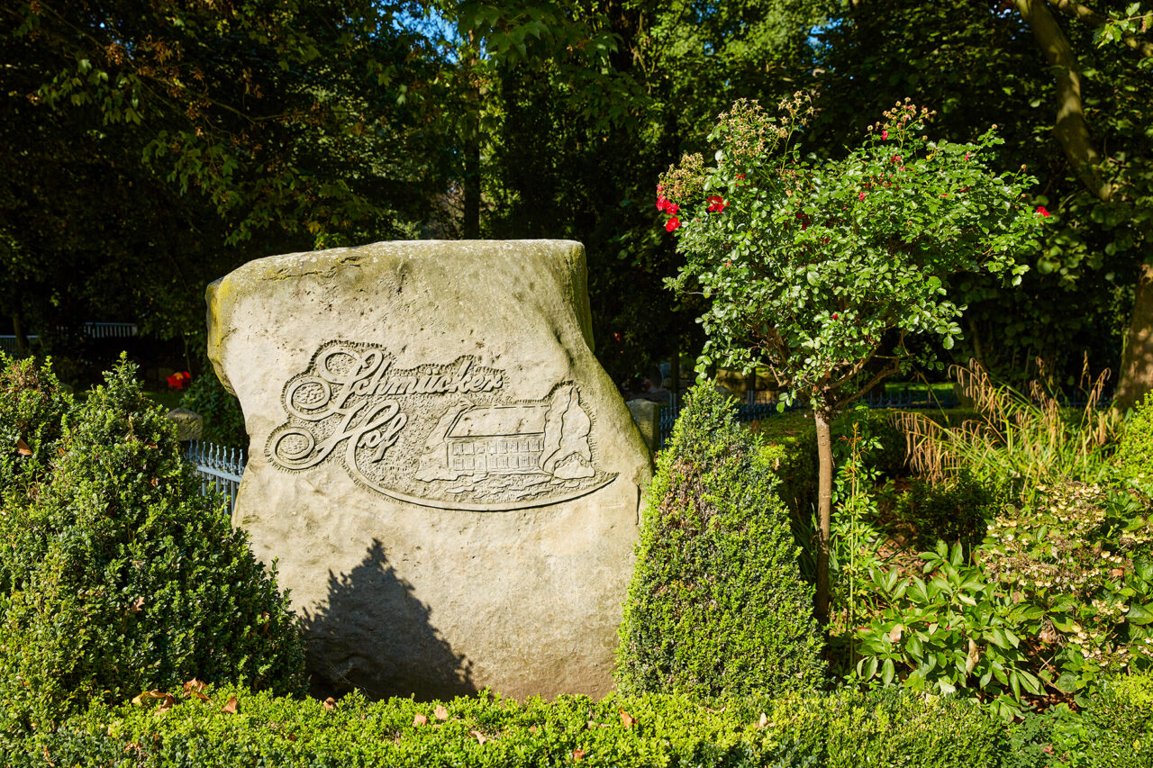 Schmücker Hof, Bottrop-Kirchhellen: Großer Stein mit "Schmachtendorf Hof" Gravur, umgeben von grünen Büschen und Bäumen in Bottrop.