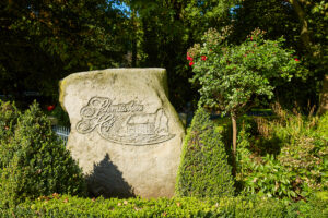 Schmücker Hof, Bottrop-Kirchhellen: Großer Stein mit "Schmachtendorf Hof" Gravur, umgeben von grünen Büschen und Bäumen in Bottrop.