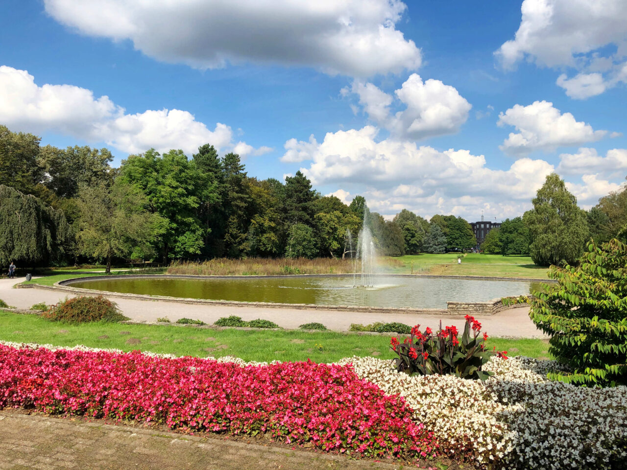 Stadtarten, Bottrop: Brunnen im Stadtgarten Bottrop mit Blumenbeeten, Wasserfläche und umgebenden Bäumen unter blauem Himmel.