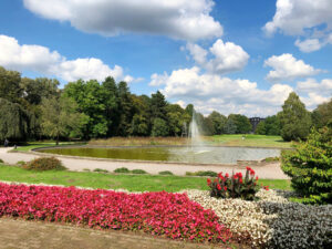 Stadtarten, Bottrop: Brunnen im Stadtgarten Bottrop mit Blumenbeeten, Wasserfläche und umgebenden Bäumen unter blauem Himmel.