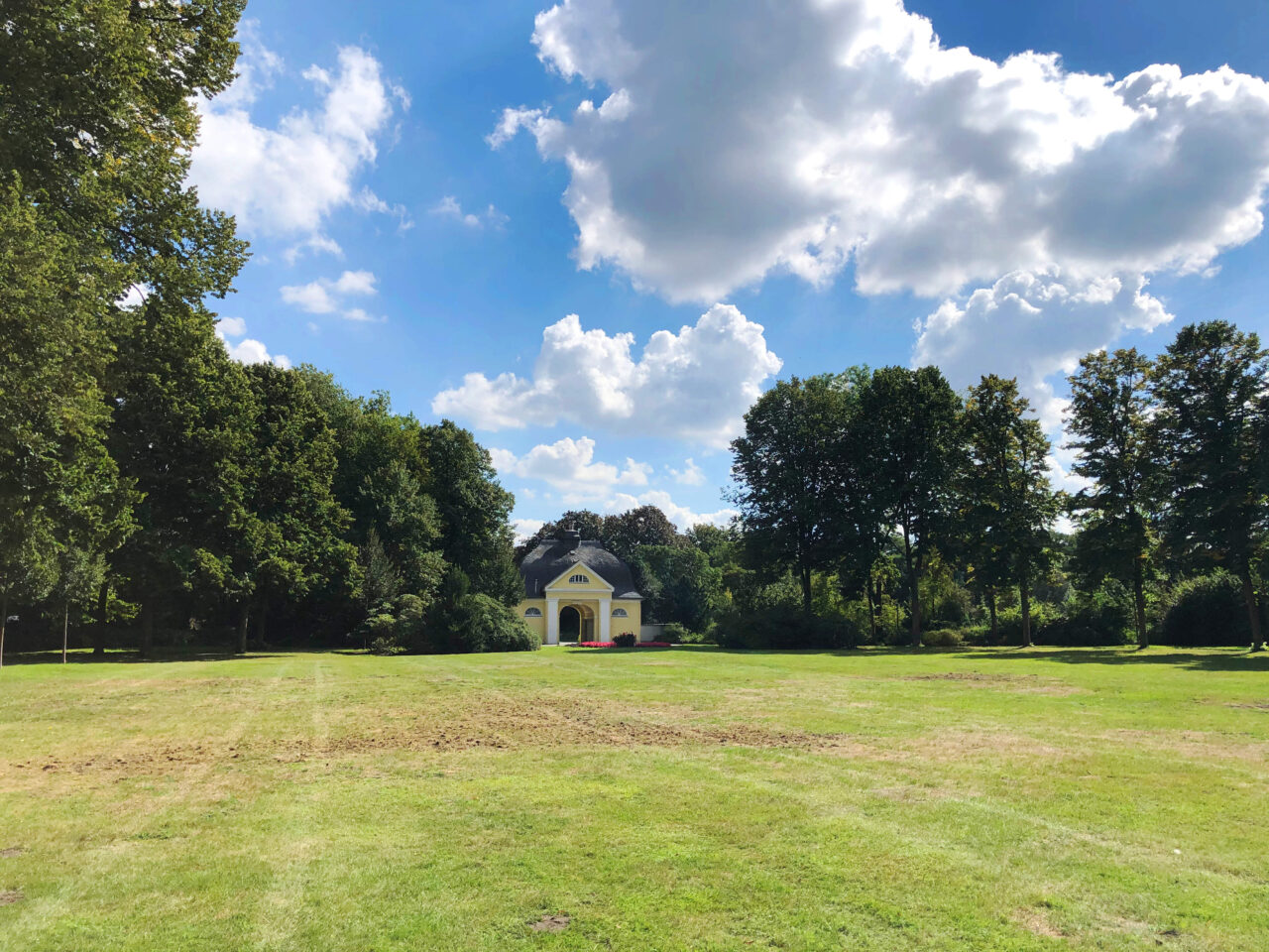 Stadtgarten, Bottrop: Grüne Parkwiese mit Bäumen und einem kleinen gelben Pavillon unter blauem Himmel mit Wolken.