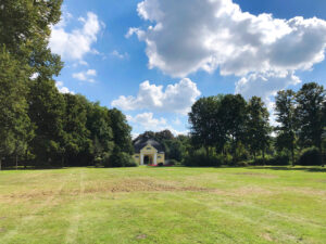 Stadtgarten, Bottrop: Grüne Parkwiese mit Bäumen und einem kleinen gelben Pavillon unter blauem Himmel mit Wolken.
