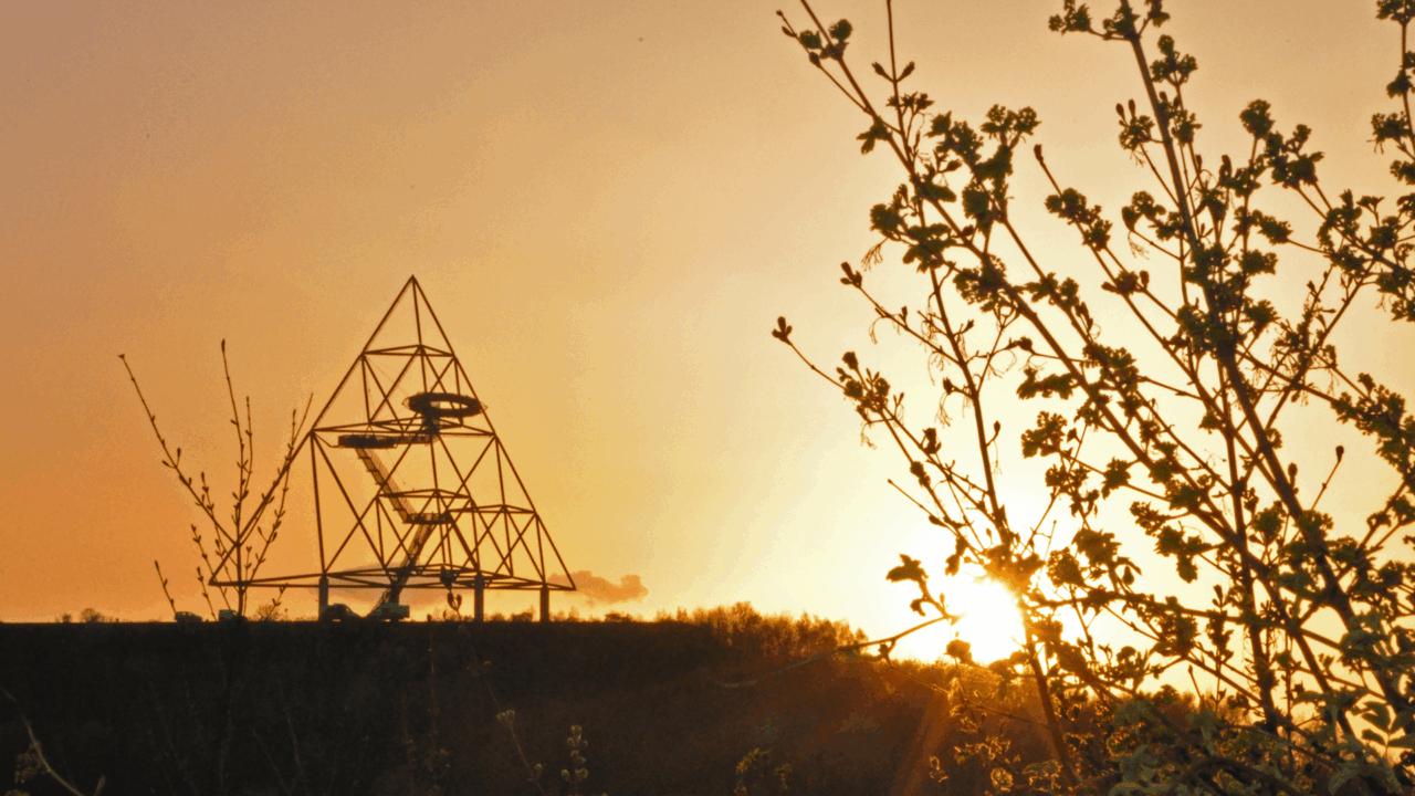 Tetraeder Bottrop: Ein eindrucksvolles Pyramidenkunstwerk bei Sonnenuntergang, mit blühenden Zweigen im Vordergrund.
