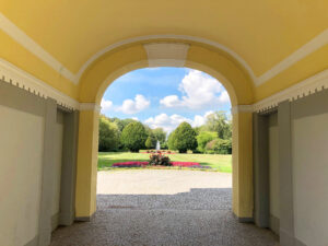 Torhaus zum Stadtgarten, Bottrop: Blick durch ein gelbes Torhaus auf den Stadtgarten Bottrop mit Rasenfläche, Blumenbeet und Springbrunnen.