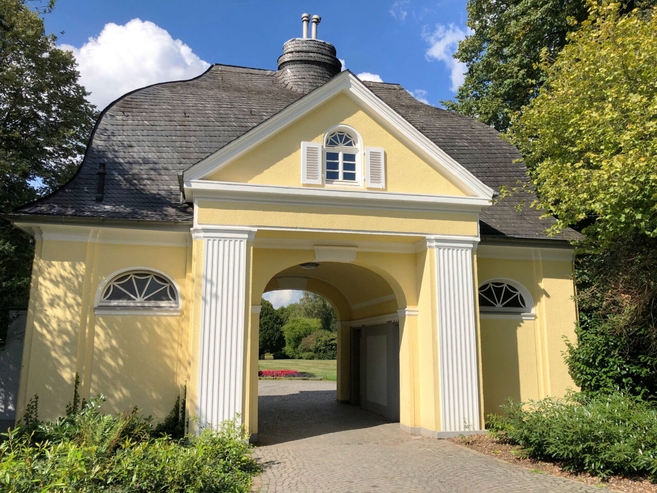 Torhaus zum Stadtgarten, Bottrop: Gelbes Torhaus mit geschwungenem Dach, weißen Säulen und Durchgang zum Stadtgarten Bottrop.