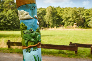 Waldkompetenzzentrum Heidhof Bottrop: Detail einer geschnitzten Holzsäule mit Frosch vor einem Spielplatz in grüner Landschaft in Bottrop.