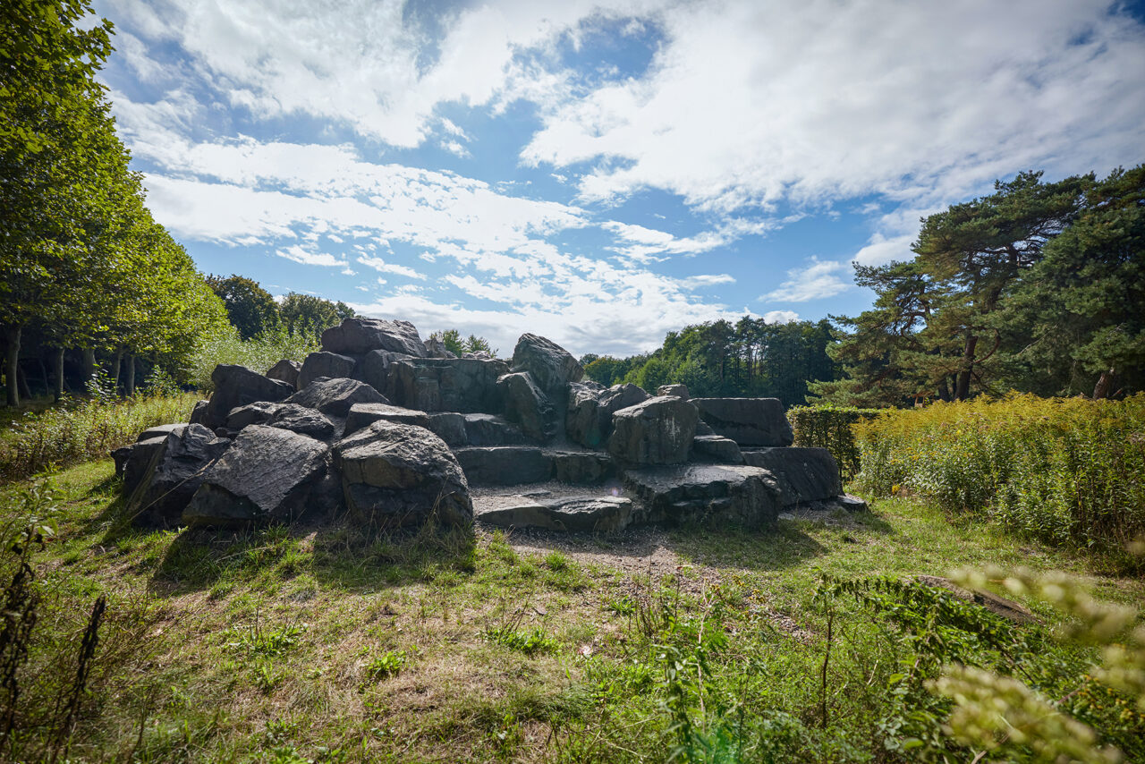 Waldkompetenzzentrum Heidhof Bottrop: ```html
Große dunkle Steine bilden einen natürlichen Felsenhügel unter einem blau-weißen Himmel.
```