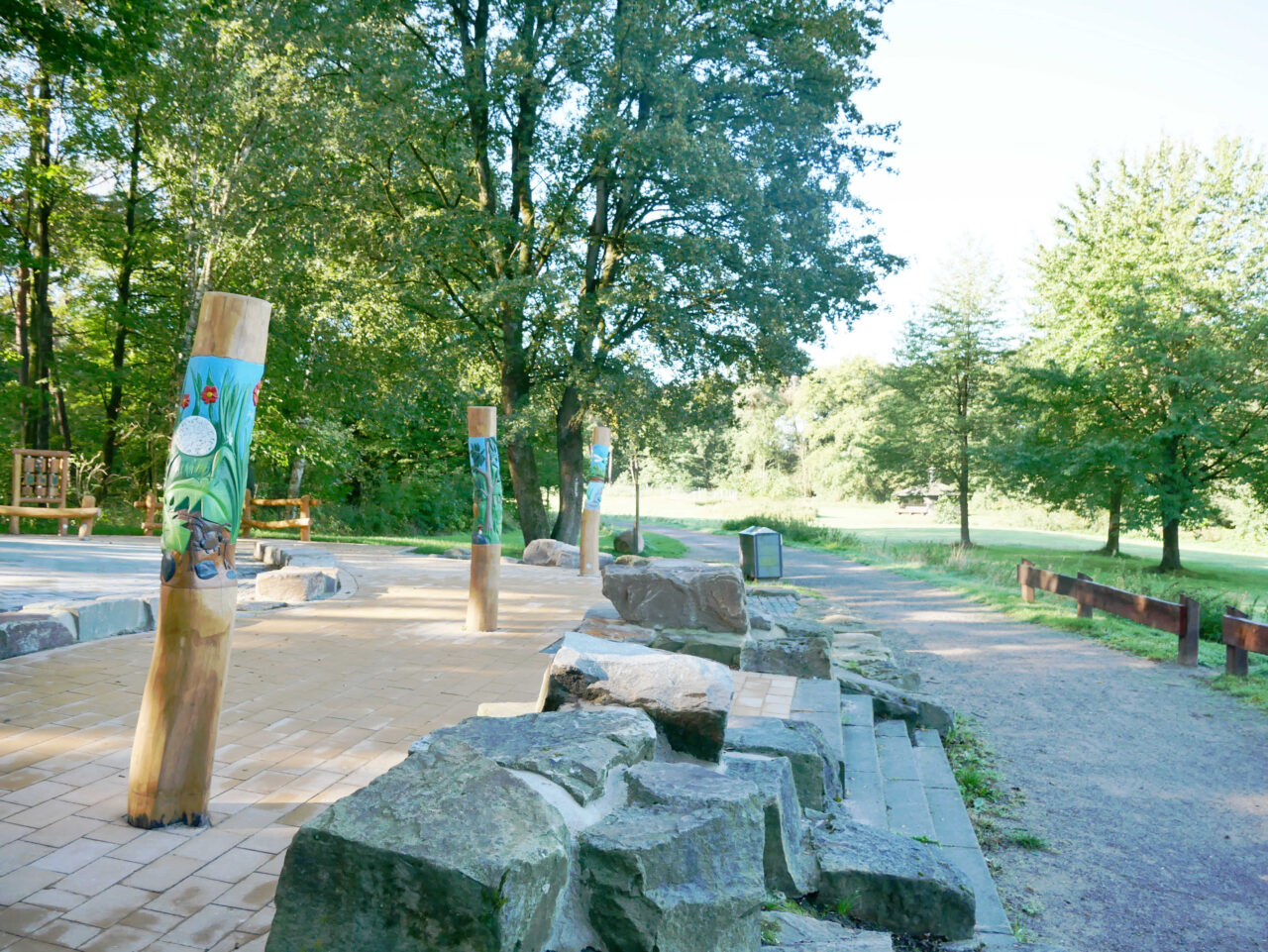 Waldkompetenzzentrum Heidhof, Bottrop, bemalte Pfähle: Bemalte Holzpfähle mit Naturmotiven stehen auf einem gepflasterten Platz im grünen Waldgebiet.