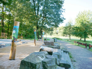 Waldkompetenzzentrum Heidhof, Bottrop, bemalte Pfähle: Bemalte Holzpfähle mit Naturmotiven stehen auf einem gepflasterten Platz im grünen Waldgebiet.